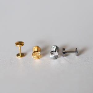 以下が含まれることがあります： 金と銀色のスタッドピアスのコレクション。ピアスは、太陽光線模様の平らな円形デザインです。ポストが見え、ピアスは白い表面に表示されています。