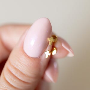 May include: Two small, gold-colored cross earrings. One earring features a small cross, while the other has a cross and a small, round charm. The earrings are held between fingers with a light pink nail polish.