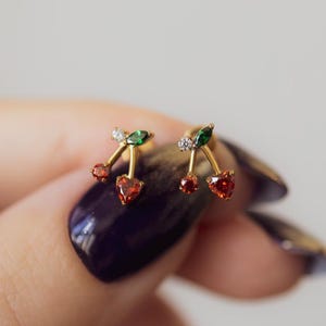 May include: A pair of gold-colored cherry stud earrings. Each earring features a red heart-shaped stone, green leaves, and small clear stones. The earrings are held up by a hand with dark purple nail polish.