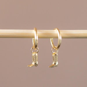 May include: A pair of gold-colored hoop earrings, each with a small, detailed cowboy boot charm. The earrings are displayed against a soft, neutral background, highlighting their delicate design and western theme.