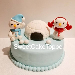 May include: A light blue cake decorated with fondant snowmen, an igloo, and snowballs. One snowman wears a blue hat and scarf, the other has red earmuffs and mittens. The cake has a scalloped edge and the text "SweetCakeTopper".