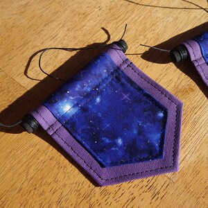 May include: Two small, pentagon-shaped fabric banners with a deep blue galaxy print and purple trim. Each banner has a black wooden dowel at the top and a thin black string for hanging. The banners are on a wooden surface.