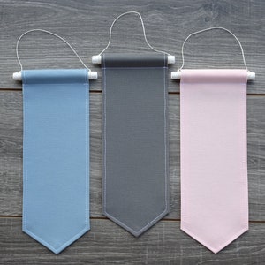 May include: Three fabric pennants hanging from white string. The pennants are light blue, gray, and pink. Each pennant has a white stitched border and a pointed bottom.