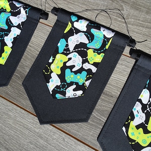 May include: Three black fabric pennants with a white and green pattern of video game controllers. The pennants are hanging from a string.