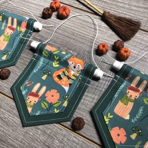May include: A green fabric banner with a fox, bunny, and mushroom pattern. The banner is strung with white twine and has white spools at each end.