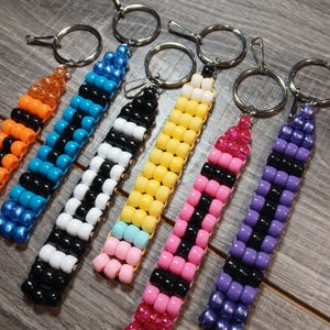 May include: A collection of beaded keychains in various colors, including orange, blue, black, white, yellow, pink, and purple. Each keychain features a beaded design with a silver keyring and clasp.