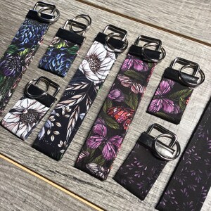May include: A set of eight keychains with different floral and butterfly designs. The keychains are made of black fabric with colorful floral and butterfly patterns. The keychains are attached to silver key rings.