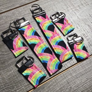 May include: Five rainbow patterned keychains with silver key rings. The keychains are different sizes and are arranged in a row on a wooden surface.