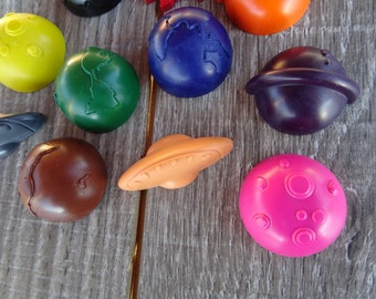 May include: A collection of colorful, planet-shaped crayons, including blue Earth, a purple planet with rings, and a pink planet with craters. Also includes a tan UFO and a yellow moon crayon.