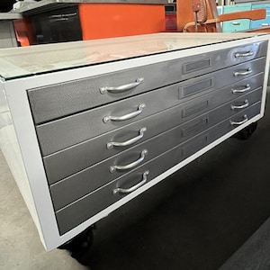 May include: A white metal file cabinet with five drawers on wheels. The drawers are gray and have silver handles. The top of the cabinet has a glass top.