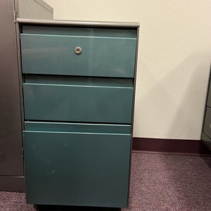 May include: A teal metal file cabinet with three drawers. The top drawer has a keyhole.