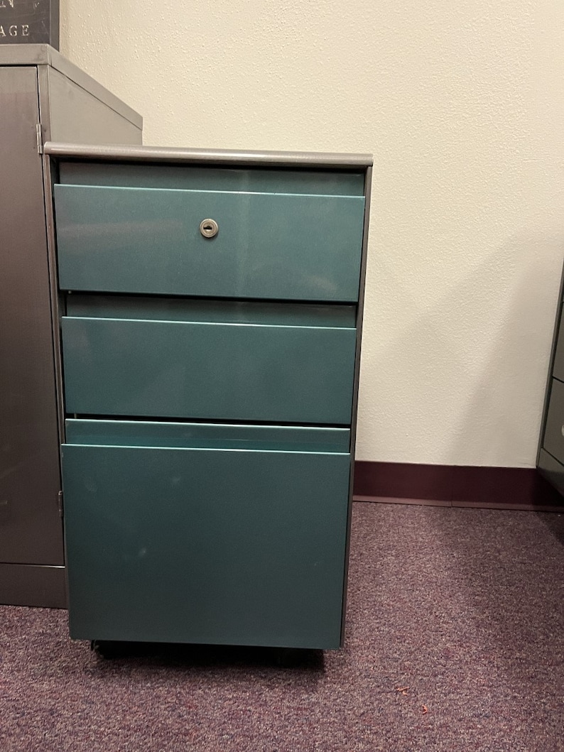 May include: A teal metal file cabinet with three drawers. The top drawer has a keyhole.