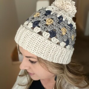 May include: A white crocheted beanie with a pom pom on top. The beanie has a multi-coloured pattern of grey, blue, and yellow squares.