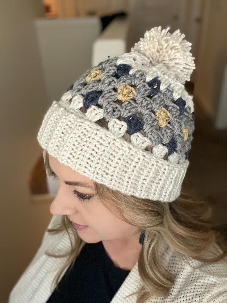 May include: A white crocheted beanie with a pom pom on top. The beanie has a multi-coloured pattern of grey, blue, and yellow squares.