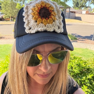 May include: A black trucker hat featuring a square, cream-colored crochet patch on the front panel. The patch displays a central sunflower motif with a dark brown center and golden-yellow petals, surrounded by a textured, off-white yarn border.