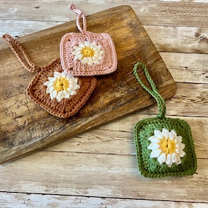 May include: Three crocheted flower ornaments with white centers and different colored petals: pink, brown, and green. The ornaments are hanging from a wooden surface.