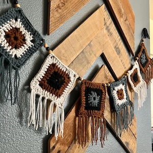 May include: A handmade crochet garland featuring square motifs in shades of brown, white, and gray, with fringe detailing. The garland is strung with wooden beads and hangs against a wooden chevron-patterned backdrop.