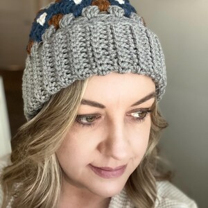 May include: A grey crocheted beanie with a colourful geometric pattern on the top. The pattern features brown, blue, white and cream squares.