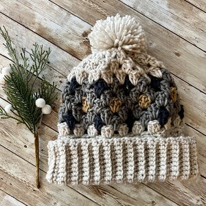 May include: A crocheted beanie hat with a pom pom on top. The hat is made in a variety of colours, including beige, grey, and brown. The hat is sitting on a wooden surface.