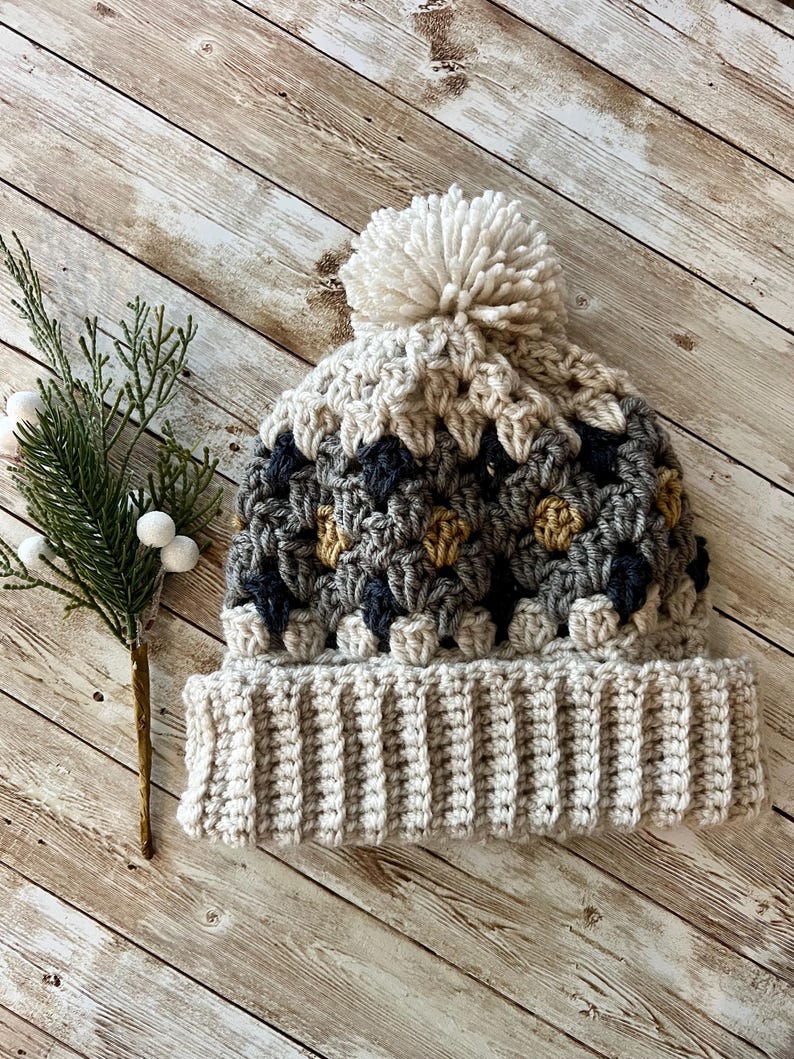 May include: A crocheted beanie hat with a pom pom on top. The hat is made in a variety of colours, including beige, grey, and brown. The hat is sitting on a wooden surface.