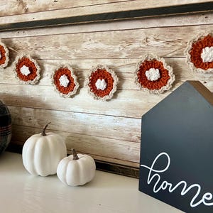 May include: A fall-themed display featuring a garland of crocheted pie slices with orange and white details. Two white pumpkins and a plaid pumpkin sit in front of a wooden sign that says "home".