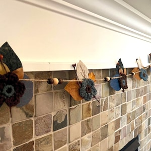 May include: A decorative garland featuring felt and crochet floral elements. The garland includes blue, brown, and yellow felt leaves and flowers, along with wooden beads strung on a natural twine. The garland is displayed against a neutral background.
