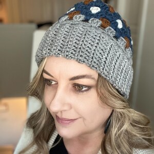 May include: A grey crocheted beanie with a colourful geometric pattern on the top. The pattern features squares of blue, brown, white, and grey yarn.