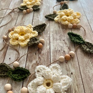 May include: A handmade crochet garland featuring cream, yellow, and green flowers, interspersed with wooden beads. The flowers have detailed petals and are connected by a natural twine. The garland is displayed on a wooden surface.