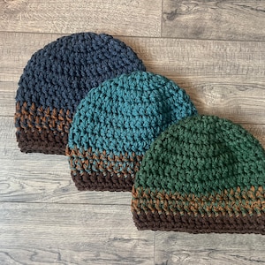 May include: Three crocheted hats in different colors: dark blue, teal, and forest green. Each hat has a brown and orange stripe around the bottom.