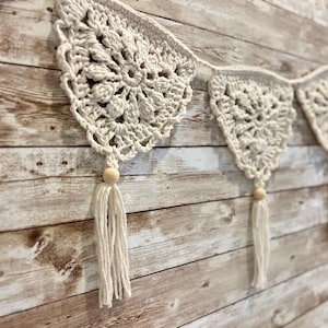 May include: A white crocheted banner with wooden beads and tassels hanging on a wood panel wall.