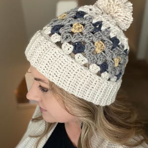May include: A white crocheted beanie with a pom pom on top. The beanie has a pattern of grey, blue, and yellow squares.