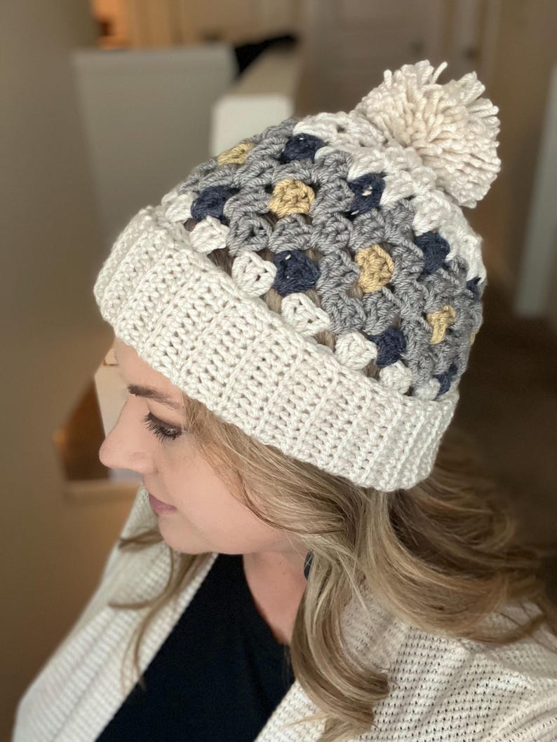 May include: A white crocheted beanie with a pom pom on top. The beanie has a pattern of grey, blue, and yellow squares.