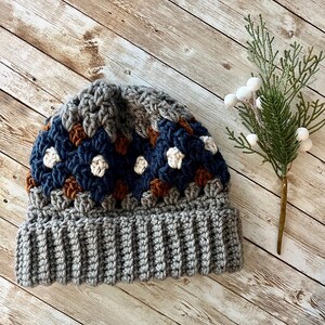 May include: A grey and blue crocheted beanie with a brown and white pattern. The beanie has a ribbed brim.