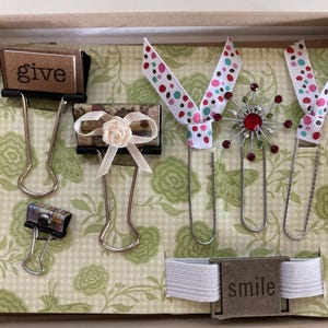 May include: A collection of decorative paper clips and accessories arranged in a box. Includes clips with ribbon, a floral embellishment, and the words "give" and "smile". The background features a green floral pattern.