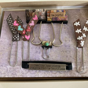 May include: A decorative box with various stationery items. Includes ribbon bookmarks with polka dots, hearts, and floral patterns. Also features binder clips, a black elastic band with the text "reality has limits, imagination is boundless," and small block letters.