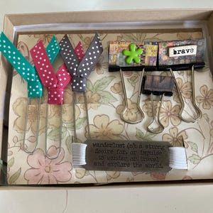 May include: A decorative box contains colorful ribbon bookmarks, binder clips, and a metal tag with the definition of wanderlust. The bookmarks have teal, pink, and gray polka dots. The word "brave" is also visible.