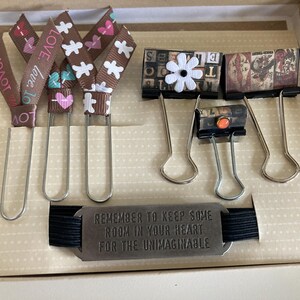 May include: A set of decorative stationery items, including ribbon bookmarks with hearts and floral patterns, binder clips, and a metal tag with the text "REMEMBER TO KEEP SOME ROOM IN YOUR HEART FOR THE UNIMAGINABLE."
