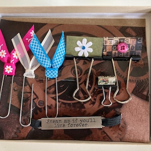 May include: A decorative box featuring handmade bookmarks and paper clips. The bookmarks have pink, white, and blue ribbons with floral and geometric patterns. The box also includes a metal tag that reads "dream as if you'll live forever."