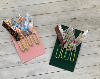 May include: Two sets of colorful ribbon bookmarks with green metal binder clips. The bookmarks are on pink and green cardstock.