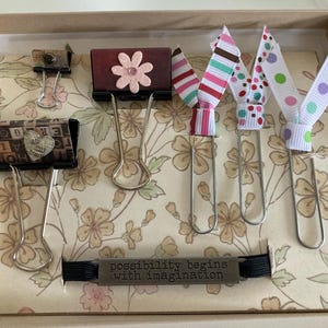 May include: A decorative box contains several binder clips and ribbon bookmarks. The binder clips are adorned with floral and letter designs. The bookmarks feature striped and polka-dot ribbons. The phrase "possibility begins with imagination" is on a metal tag.