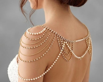 May include: Gold-coloured shoulder jewellery featuring multiple strands of faux pearls and clear crystals. The shoulder piece drapes over the shoulders and back. The jewellery is worn over a white dress.