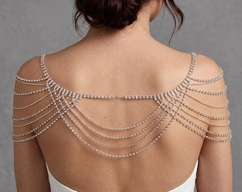 May include: A silver shoulder necklace with multiple strands of small, sparkling rhinestones. The necklace drapes over the shoulders and back, creating a delicate and elegant look. The woman is wearing a white dress.