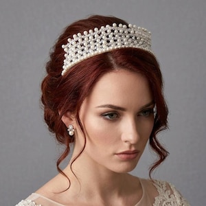 May include: A bridal tiara with a delicate design, featuring numerous small, white pearls. The tiara sits atop a person's auburn hair, complemented by matching pearl earrings. The background is a soft gray.