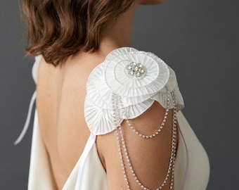 May include: White shoulder accessory with layered, pleated fabric and a central rhinestone embellishment. Delicate, cascading rhinestone chains hang from the shoulder piece. The accessory is designed to be worn over a white dress.