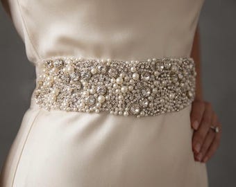 May include: A wide, decorative belt featuring an intricate arrangement of white pearls and clear, faceted crystals. The embellishments are densely packed across the entire surface of the belt, creating a textured, shimmering appearance.