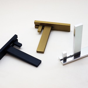 May include: Three modern shelf brackets in black, gold, and silver. The brackets have a minimalist design with a horizontal base and a vertical support. The black bracket is on the left, the gold in the middle, and the silver on the right.