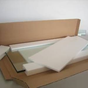 May include: A large, rectangular piece of glass is carefully packaged in a cardboard box with white foam padding. The glass is clear and appears to be a mirror or window pane.