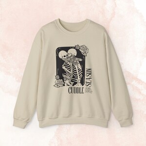 Puede incluir: Sudadera color crema con un gráfico negro de dos esqueletos abrazándose. El diseño incluye las palabras "Cuddle Season" e ilustraciones de rosas. Prenda de vestir informal.