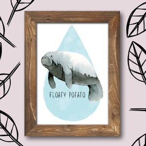May include: A framed watercolour print featuring a manatee. The manatee is depicted in shades of grey and white, set against a light blue backdrop. The text "FLOATY POTATO" is below the manatee. The background is a pale pink with black leaf illustrations.