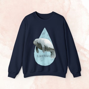 Puede incluir: Sudadera azul marino con una ilustración de acuarela de un manatí dentro de una forma de lágrima azul claro. El texto "FLOATY POTATO" está impreso debajo. Sudadera de estilo casual.